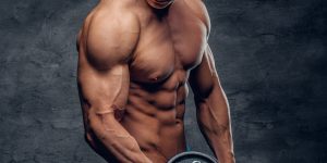 Athletic shirtless young male fitness model holds the dumbbell with light isolated on dark background.
